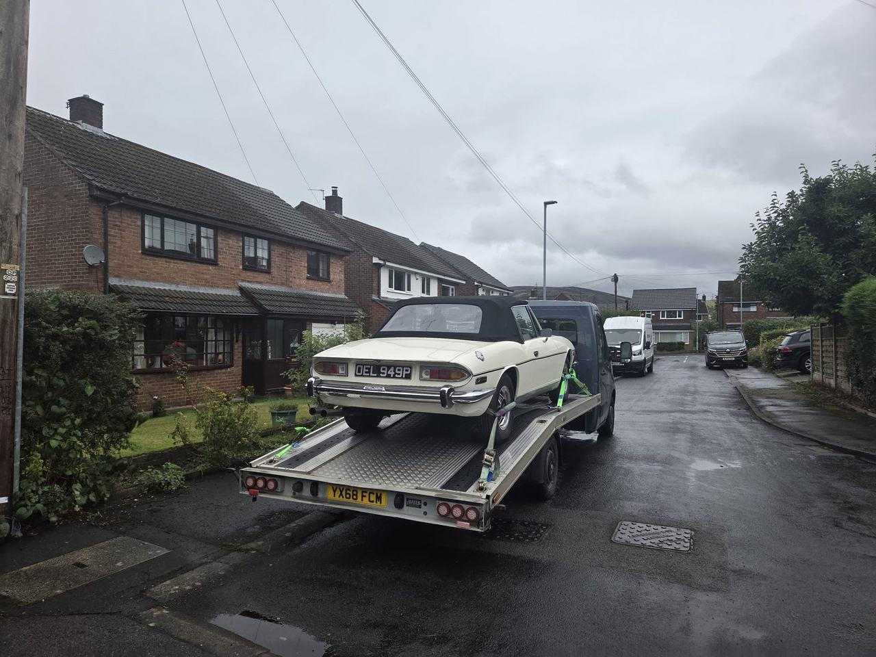 Triumph White Stag with a black soft top, strapped to a flatbed transporter on a wet residential street under a grey sky.