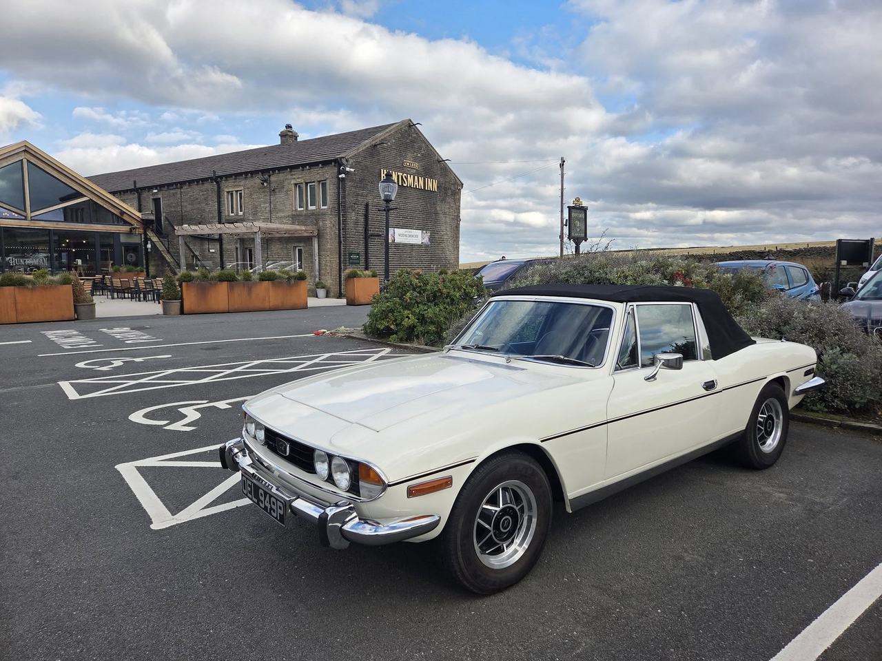Triumph White Stag with a black soft top parked in front of the stone Huntsman Inn, under a cloudy sky.