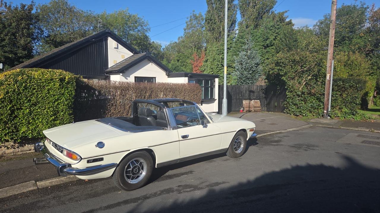 Triumph White Stag with the soft top down, parked in sunshine beside a hedge.